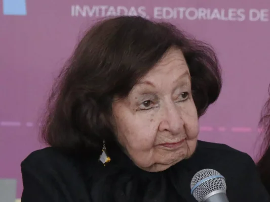 Amparo Dávila speaking into a microphone at a conference against a vibrant pink background with partial text behind her.