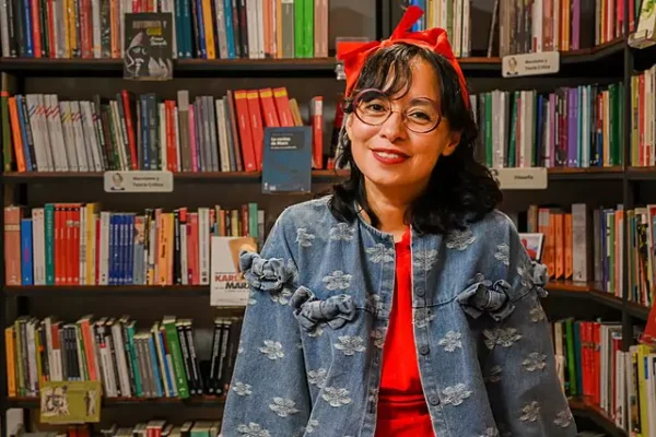 Brenda Navarro in a denim floral jacket, standing in front of a bookshelf filled with diverse books, representing her role as a contemporary Mexican author.