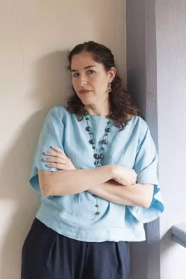 Guadalupe Nettel standing with arms crossed, wearing a light blue blouse and dark pants, leaning against a wall with a calm, introspective expression.