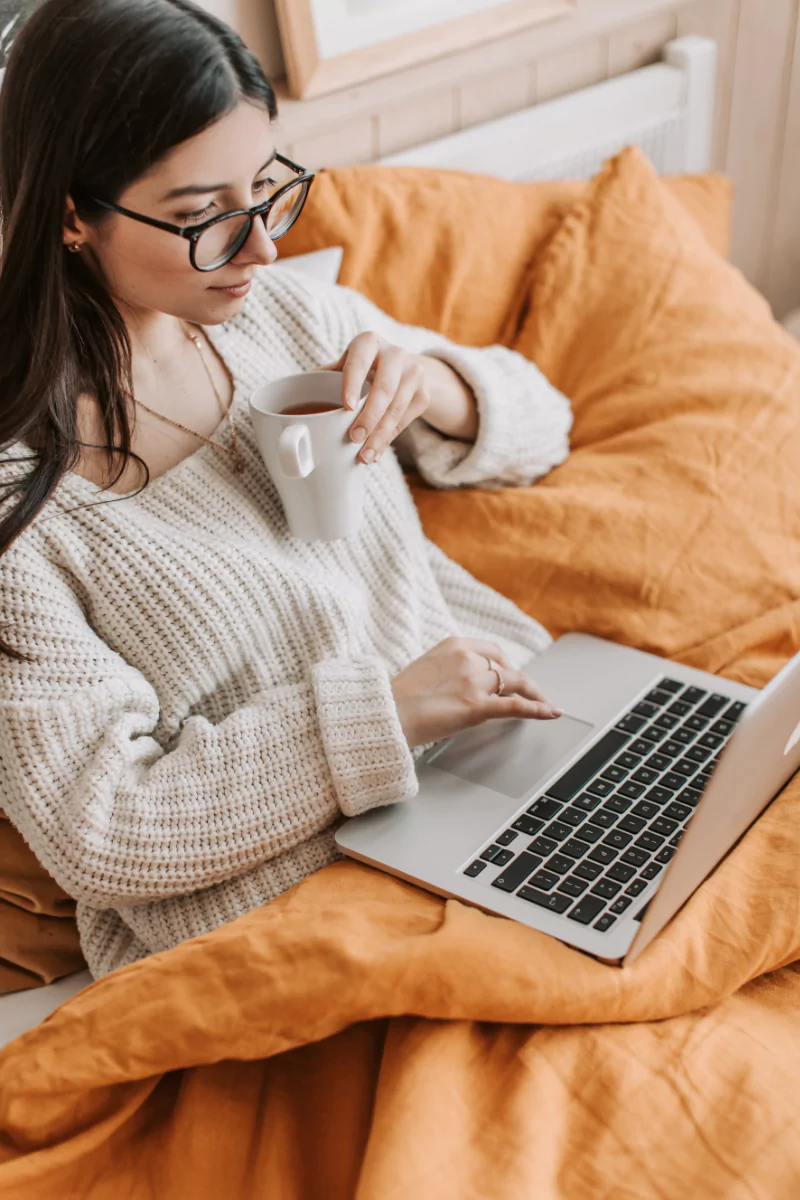 Woman enjoying coffee while working on a laptop in a cozy bedroom setting.